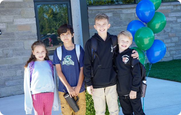 Students standing in front of blue and green balloons with their backpacks outside the school