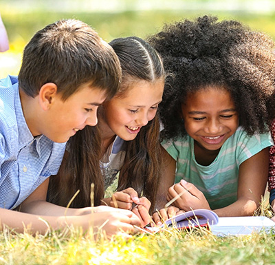 Three students smiling and writing together outdoors on the grass