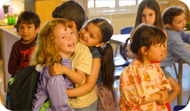 Group of children gathered together in a classroom, with two children embracing in the foreground.