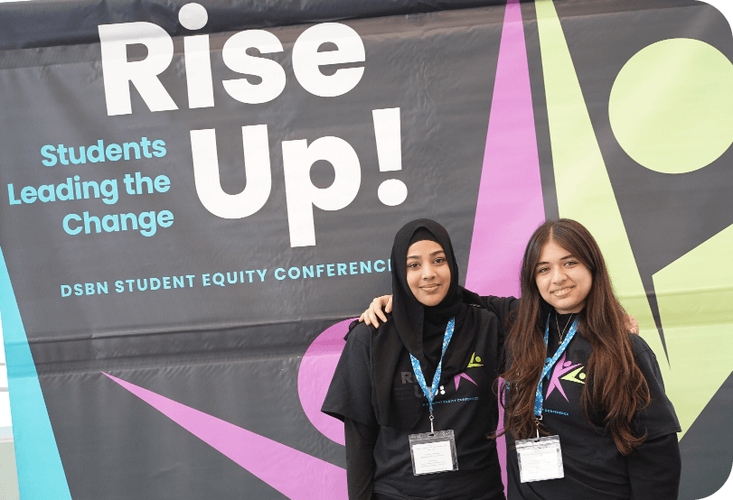 Two students standing in front of a banner for the DSBN Student Equity Conference that reads “Rise Up! Students Leading the Change.”