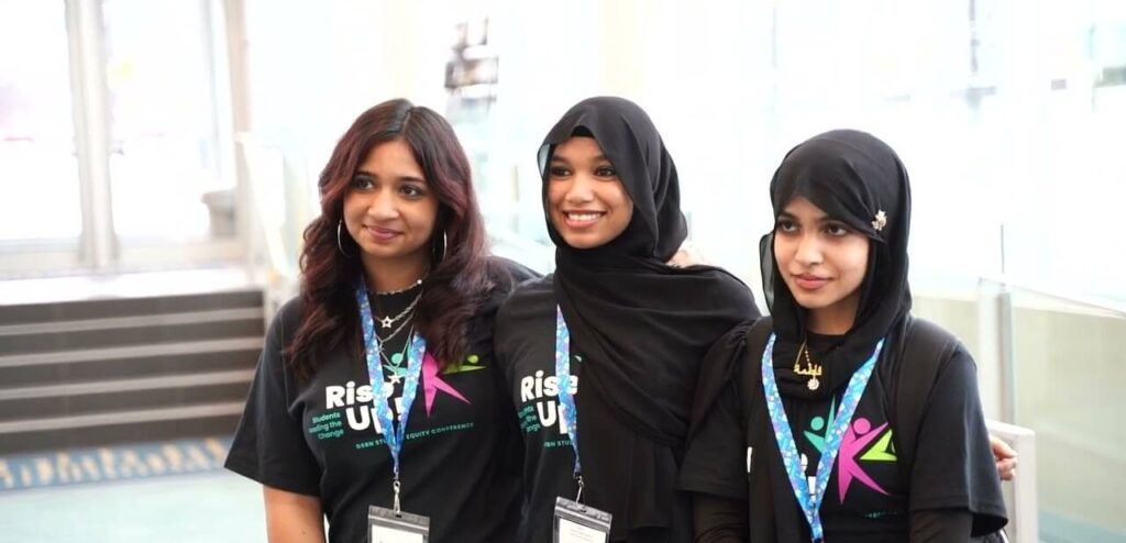 Three students wearing conference lanyards and Rise Up event shirts, posing together at the DSBN Student Equity Conference.