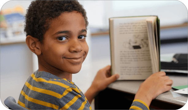 Student reading a book while seated at a classroom desk.