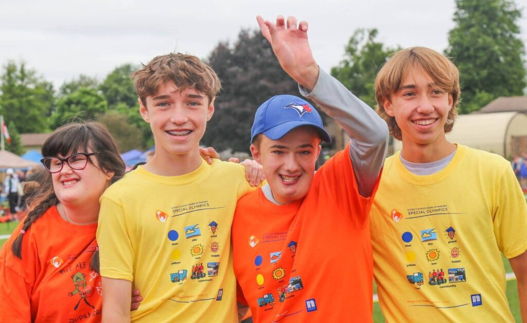 Four students wearing Special Olympics event T-shirts, standing together on an outdoor athletic field.