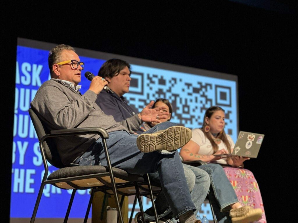Panel discussion featuring speakers seated on a stage, with one speaker using a microphone and a presentation screen visible in the background.