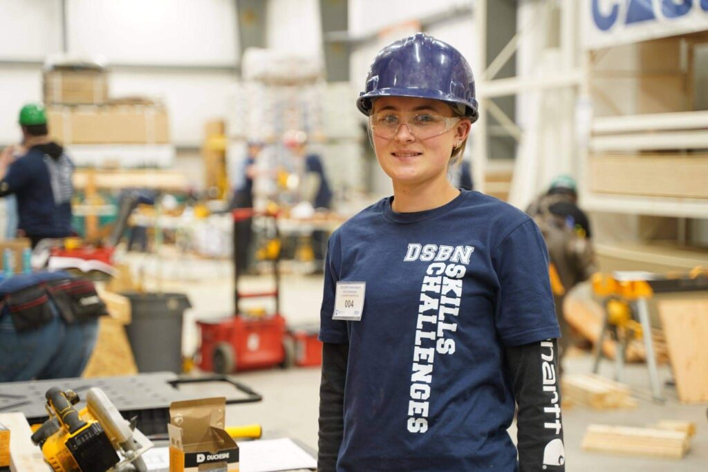 Student wearing protective equipment in a skilled trades workshop, standing among tools and construction materials.