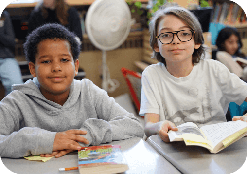 Two students seated at a classroom table with books in front of them.